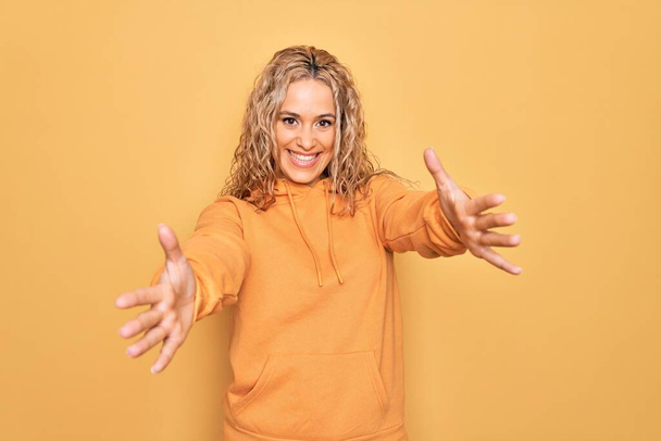 Junge schöne blonde, sportliche Frau in lässigem Sweatshirt vor gelbem Hintergrund, die lächelnd mit offenen Armen in die Kamera blickt, um sich zu umarmen. Fröhlicher Ausdruck des Glücks. - Foto, Bild