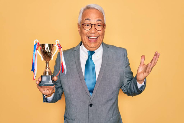 Hombre de negocios de mediana edad de cabello gris con gafas que sostienen el trofeo ganador muy feliz y emocionado, expresión ganadora que celebra la victoria gritando con una gran sonrisa y levantando las manos
 - Foto, imagen