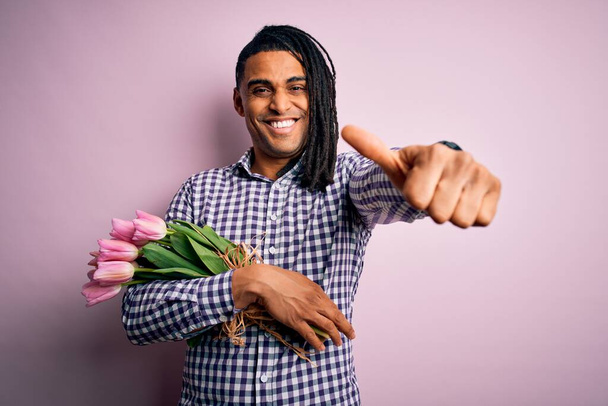 Mladý afro-romantik z Afriky s dredy v ruce drží kytici růžových tulipánů, schvaluje pozitivní gesto rukou, palce nahoru, usmívá se a raduje se z úspěchu. Gesto vítěze. - Fotografie, Obrázek