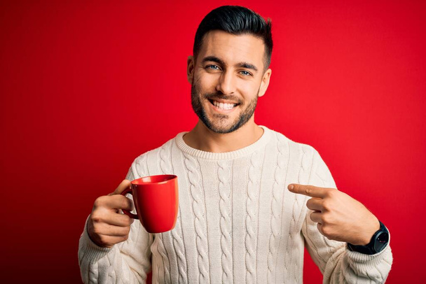 Young handsome man drinking a cup of hot coffee over red isolated background with surprise face pointing finger to himself - Foto, imagen