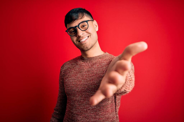Joven hombre hispano guapo con gafas nerd sobre fondo rojo sonriente alegre ofreciendo mano de palma dando asistencia y aceptación
. - Foto, Imagen