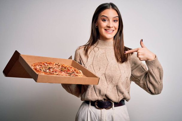 jong mooi brunette vrouw holding box met italiaanse pizza over witte achtergrond met verrassing gezicht wijzend vinger naar zichzelf - Foto, afbeelding