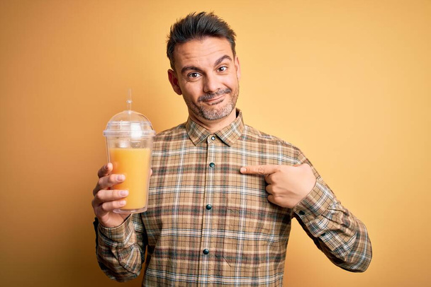 Молодой красивый мужчина пьет здоровый апельсиновый сок с помощью соломы на желтом фоне с удивленным лицом, указывающим пальцем на себя
 - Фото, изображение