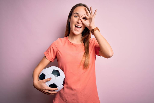 Młoda piękna gracz kobieta gra w piłkę nożną trzymając piłkę nożną na różowym tle z uśmiechem szczęśliwej twarzy robi ok znak z ręki na oko patrząc przez palce - Zdjęcie, obraz