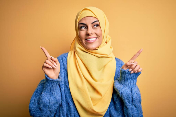 Genç, güzel, esmer, Müslüman bir kadın izole edilmiş sarı arka plan üzerine Arap başörtüsü takıyor ve parmaklarıyla farklı yönleri işaret ederek gülümsüyor. Reklam için boşluk kopyala - Fotoğraf, Görsel
