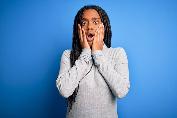 Junge afrikanisch-amerikanische Frau steht mit lässigem Rollkragen über blauem isoliertem Hintergrund und ist verängstigt und schockiert, überrascht und erstaunt mit den Händen im Gesicht - Foto, Bild