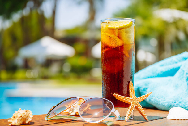 Ποτήρι cuba libre cocktail με κοχύλια, γυαλιά ηλίου και μπλε πετσέτα - Φωτογραφία, εικόνα