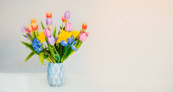 Fresh spring pastel colors bouquet of tulips, daffodils, irises in vase standing on white surface with light beige background. Festive flowers for gift. Mockup for greeting card. Wide banner - Photo, image