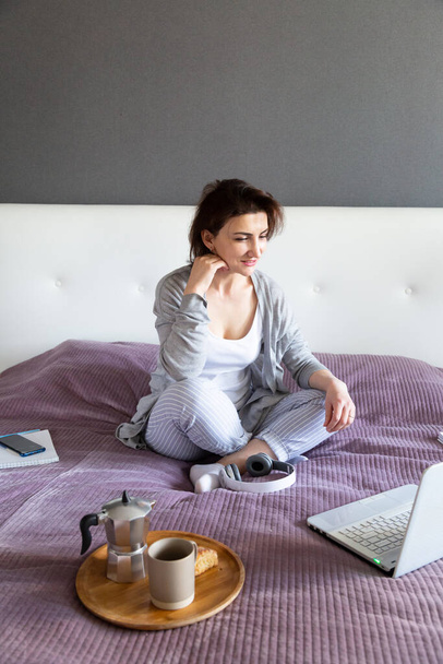 Jonge vrouw in grijs werken thuis in haar slaapkamer, freelance werk met laptop. - Foto, afbeelding
