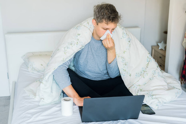 Young caucasian sick man blowing his nose sitting on the bed under a warm blanket and working on a laptop. Work at home during an illness. Self isolating. Copy space, selective focus. - Фото, зображення