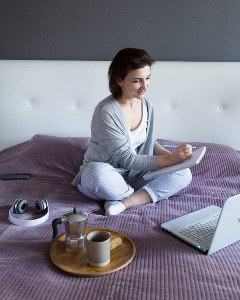 Junge Frau in Grau arbeitet zu Hause im Schlafzimmer, freiberuflich mit Laptop. - Foto, Bild
