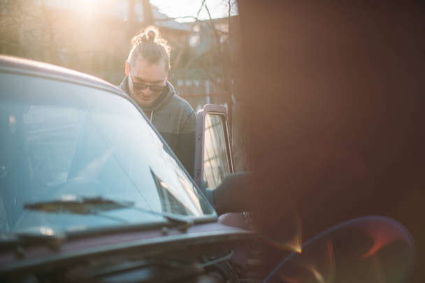 Ένας άντρας επισκευάζει ένα αυτοκίνητο, ανοίγει το καπό. Ένας νεαρός κοιτάζει κάτω από το καπό ενός αυτοκινήτου, αυτο-επισκευή σε ένα ιδιωτικό γκαράζ - Φωτογραφία, εικόνα