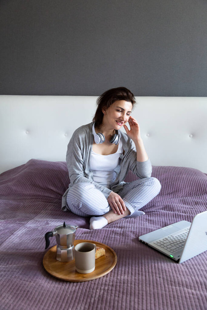 Χαμογελαστή γυναίκα σε γκρι χαλαρώνει στο σπίτι, κάθεται στο κρεβάτι, βλέποντας μια ταινία στο φορητό υπολογιστή με ακουστικά, ακούγοντας μουσική. - Φωτογραφία, εικόνα