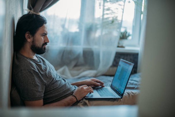 Een jongeman zit op een bank en werkt thuis op een laptop. De in quarantaine geplaatste persoon werkt vanuit huis. Werk op afstand, thuiswerk. - Foto, afbeelding