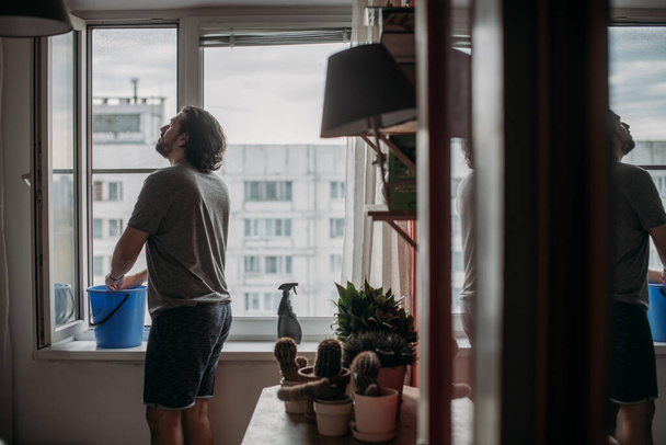 Un uomo lava le finestre a casa. Giovane bel ragazzo che fa le pulizie di primavera in un appartamento accogliente. - Foto, immagini