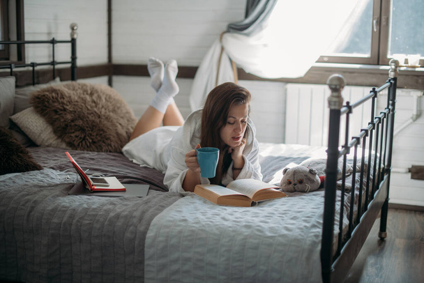 女性が本を持ってベッドに横になっている。若い女の子休みます,午後に大きな居心地の良いベッドの上の寝室で本を読んで - 写真・画像
