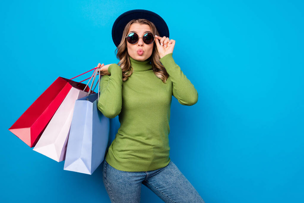Photo of cheerful funny curly lady hold many shop bags packs shopaholic send air kisses wear sun specs hat casual green turtleneck jeans isolated blue color background - Photo, image