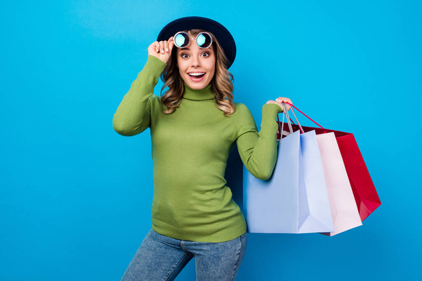 Photo of cheerful funny curly lady hold many shop bags packs addicted shopaholic excited mood wear sun specs hat casual green turtleneck jeans isolated blue color background - 写真・画像