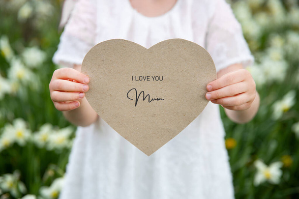 Je t'aime maman écrite sur une carte en forme de coeur tenue par une fille sur fond de fleur et d'herbe
 - Photo, image