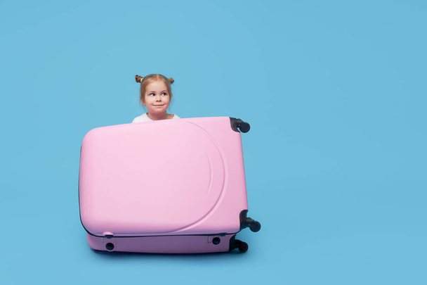 Cute baby girl sitting in a suitcase and waiting for summer vacation after quarantine. Pointing to the side. Blue studio background.  - Fotografie, Obrázek