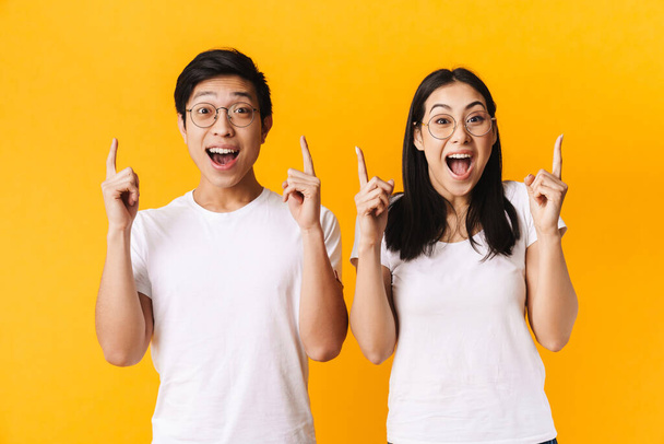 Image of delighted multinational man and woman in eyeglasses pointing finger upward isolated over yellow background - Zdjęcie, obraz