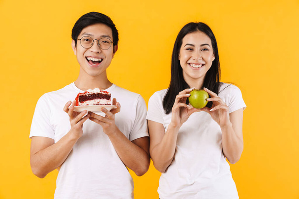 Image of smiling multinational couple holding cake and green apple isolated over yellow background - Zdjęcie, obraz