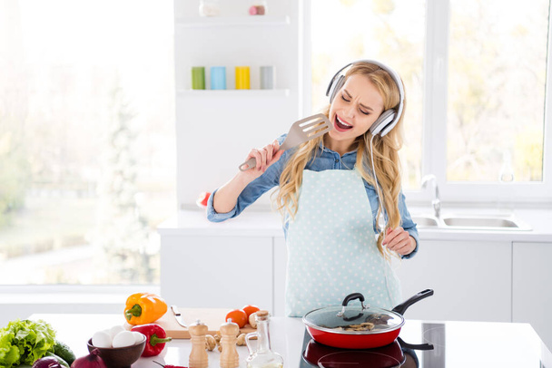 Photo of cheerful funny housewife blond lady cutting salad cooking tasty dinner breakfast listening cool headphones music sing favorite song into spoon utensil kitchen indoors - Fotografie, Obrázek