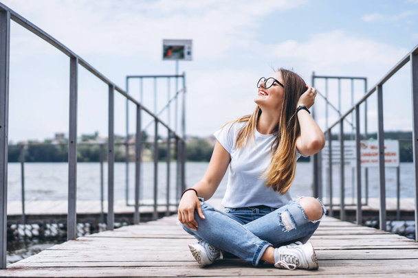 Young woman with long hair in stylish glasses posing on a wooden pier near the lake. Girl dressed in jeans and t-shirt smiling and looking away - Фото, изображение