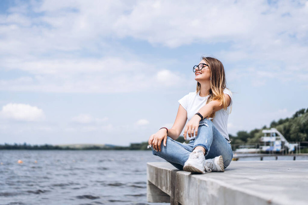 Young woman with long hair in stylish glasses posing on the concrete shore near the lake. Girl dressed in jeans and t-shirt smiling and looking away. - Photo, Image