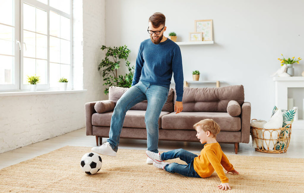 Ganzer Länge fröhlicher Junge lachend und auf dem Boden liegend neben bärtigem Vater mit Ball, während er zu Hause Fußball spielt - Foto, Bild
