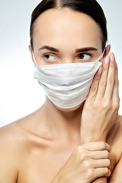 young brunette girl looking sideways wearing a white medical mask, near her face well-groomed hands clasped next  - Foto, afbeelding