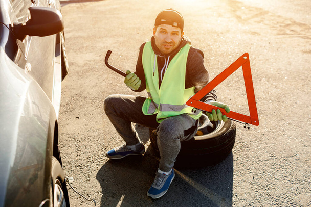 Worker or driver repairing a car. He throws up his hands and does not know what to do. Concerned and worried. Bad worker. He is dressed in a signal vest. He has a road triangle in his hands. Sits on a - Photo, Image