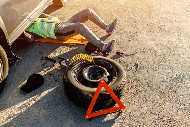 A driver or worker repairs a broken car on the side of the road. View from above. Man is under the car - Фото, изображение