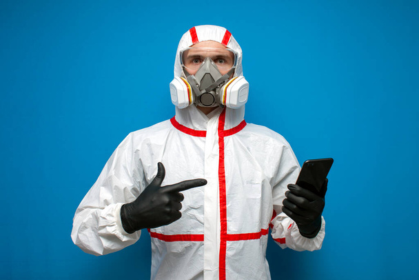 desinfectante profesional hombre en un traje de protección sostiene un teléfono inteligente y lee noticias sobre coronavirus en un fondo aislado azul
 - Foto, Imagen