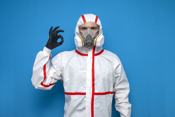 профессиональный дезинфицирующий человек в защитном костюме показывает в порядке знак на синем изолированном фоне, дезинфекционный работник, коронавирусная концепция
 - Фото, изображение