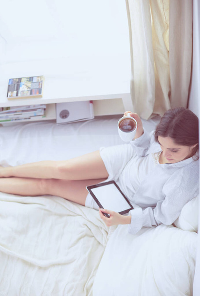 Portrait of woman using tablet and drinking coffee while sitting on the bed in the morning - Fotó, kép
