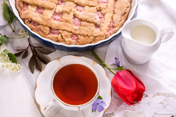 Wit porseleinen kopje thee en vers gebakken taart. Brits ontbijt stilleven met drank en lekkernijen, rode tulpenbloem en wit tafelkleed. - Foto, afbeelding
