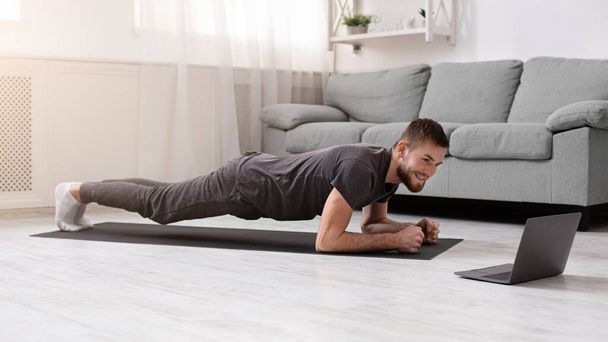 Young man doing plank exercise with online tutorial at home - 写真・画像