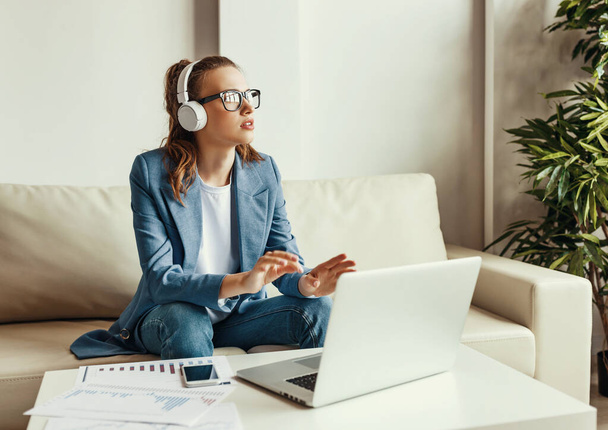 Cheerful young female in casual outfit and glasses having video conversation with colleagues and sharing business ideas while sitting on couch in modern workplac - Foto, afbeelding