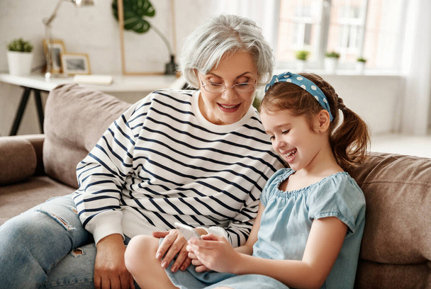 Kleines Mädchen lächelt und zeigt einer älteren Frau ihr Smartphone, während sie zu Hause auf der Couch sitzt - Foto, Bild