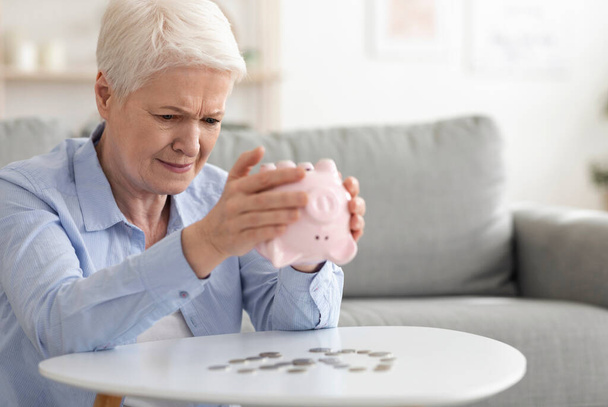 Finanzkrise. Traurige Seniorin schüttelt Sparschwein und nimmt Münzen heraus - Foto, Bild