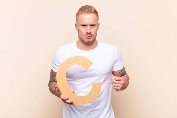 young blonde man angry, anger, disagreement, holding the letter C of the alphabet to form a word or a sentence. - Фото, изображение