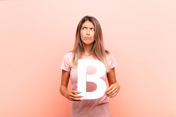 young pretty woman confused, doubtful, thinking, holding the letter B of the alphabet to form a word or a sentence. - Foto, Imagen
