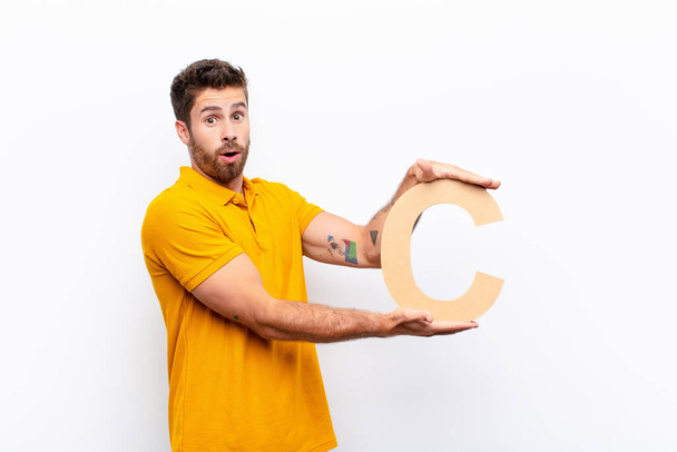 young handsome man surprised, shocked, amazed, holding the letter C of the alphabet to form a word or a sentence. - Фото, зображення