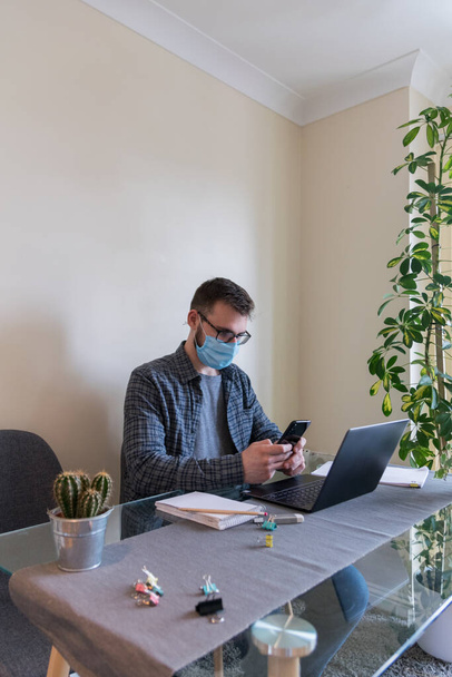 Вид сбоку на молодого европейца в маске для лица, работающего из дома из-за короно-вирусных ограничений использования ноутбука и проверки мобильного телефона
 - Фото, изображение
