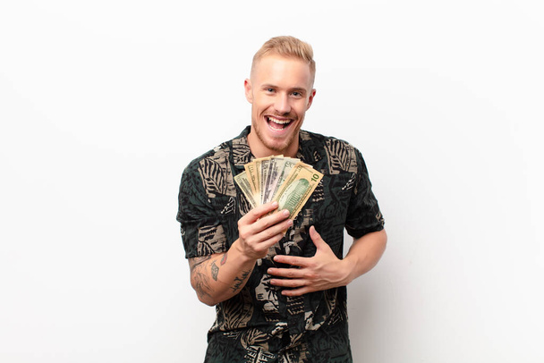 young blonde man laughing out loud at some hilarious joke, feeling happy and cheerful, having fun against white wall with banknotes - Fotoğraf, Görsel