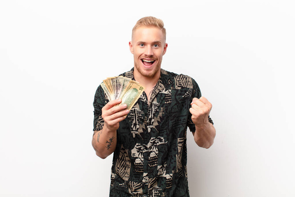 young blonde man feeling shocked, excited and happy, laughing and celebrating success, saying wow! against white wall with banknotes - Photo, image