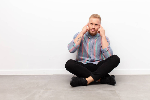 young blonde man feeling confused or doubting, concentrating on an idea, thinking hard, looking to copy space on side sitting on cement floor - Foto, imagen