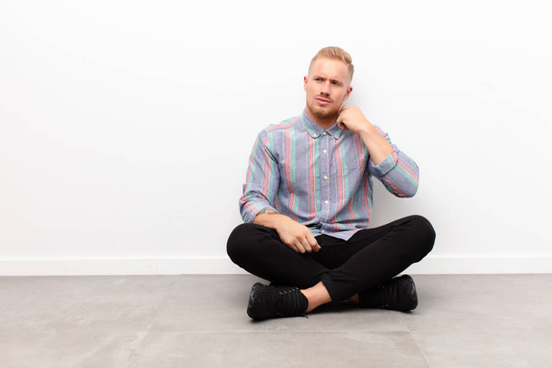 young blonde man feeling stressed, anxious, tired and frustrated, pulling shirt neck, looking frustrated with problem sitting on cement floor - Foto, Imagem