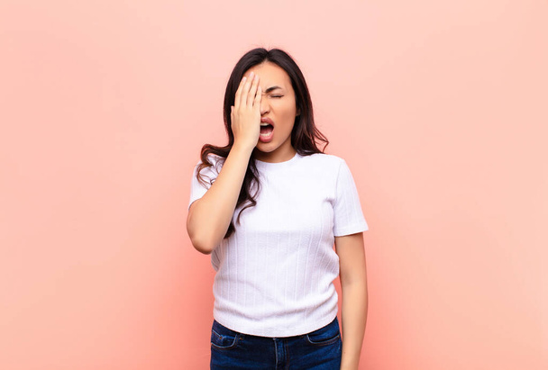 young latin pretty woman looking sleepy, bored and yawning, with a headache and one hand covering half the face against flat wall - Fotoğraf, Görsel